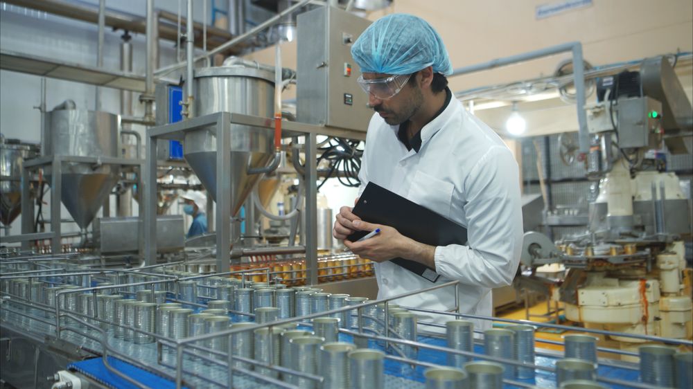 A quality officer in a lab coat, hairnet and safety glasses recording checks against product moving along a processing line